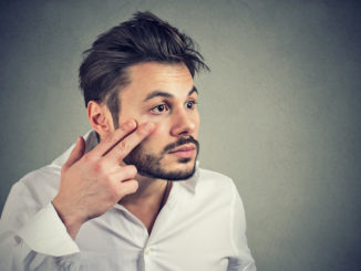 Young man pulling down eyelid checking his eye looking in mirrow feels unwell has black circles