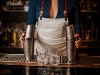 Female bartender in the white apron holding in her hands two steel cocktail shakers standing at the bar counter
