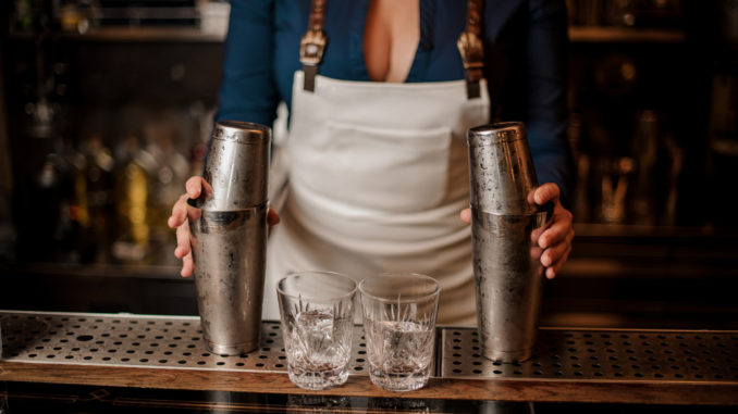 Female bartender in the white apron holding in her hands two steel cocktail shakers standing at the bar counter