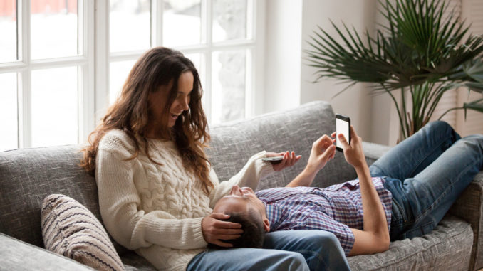 Cheerful young couple relaxing on sofa talking using smartphones, loving positive men and women laughing having fun enjoying conversation holding mobile phones at home living room on couch together