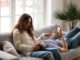 Cheerful young couple relaxing on sofa talking using smartphones, loving positive men and women laughing having fun enjoying conversation holding mobile phones at home living room on couch together