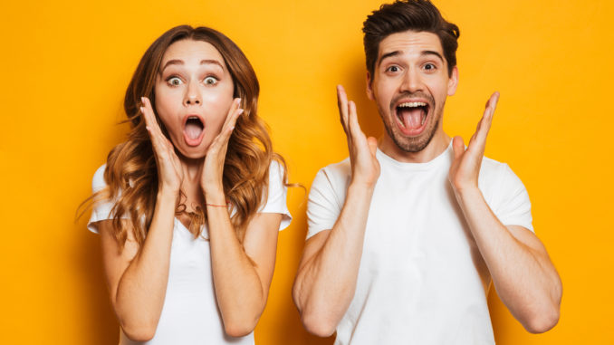 Photo of excited couple men and women in basic clothing shouting in surprise or delight and raising arms isolated over yellow background