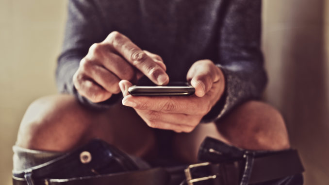 Closeup of a young caucasian man using his smartphone in the toilet while sitting in the bowl