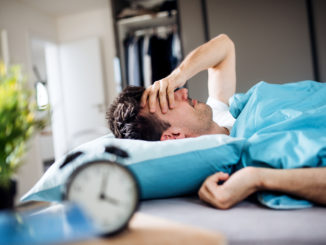 A young tired man with alarm clock in bed at home, waking up in morning.