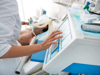 Close up of female hands touching monitor of mechanical ventilator. Middle aged man lying in hospital bed on blurred background