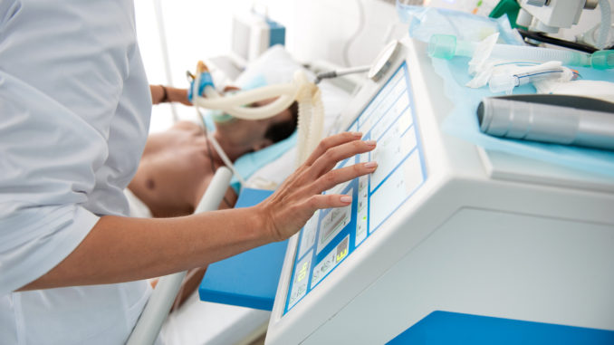 Close up of female hands touching monitor of mechanical ventilator. Middle aged man lying in hospital bed on blurred background