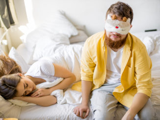 Sleepy caucasian men in mask for sleeping sit on bed near sleeping wife,in the morning at home. men want to sleep, before going to work.