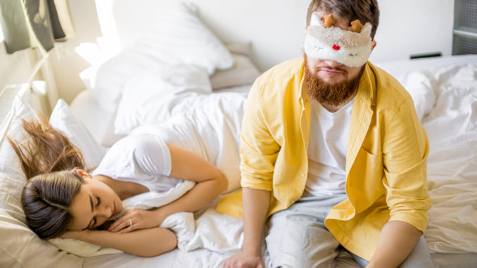 Sleepy caucasian men in mask for sleeping sit on bed near sleeping wife,in the morning at home. men want to sleep, before going to work.