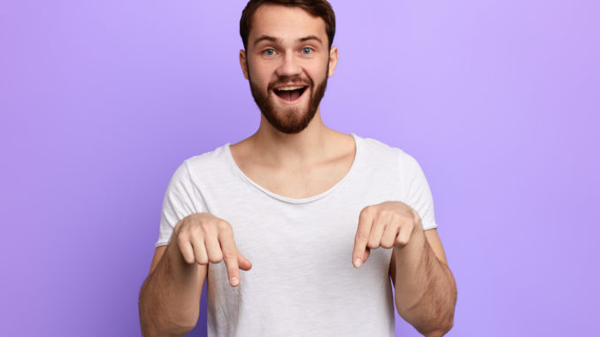 Handsome positive bearded man pointing down with two fingers.