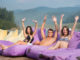 Man is surrounded by two women sitting on a cushioned lounger with his hands raised up