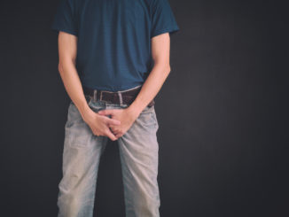 Man in jeans covers his crotch with hands against black wall.