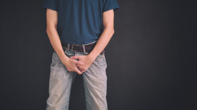 Man in jeans covers his crotch with hands against black wall.