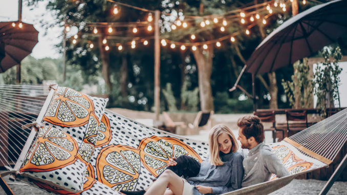 Stylish hipster couple cuddling and relaxing in hammock under retro lights in evening summer park. rustic men and women embracing and resting in forest. space for text. summer vacation