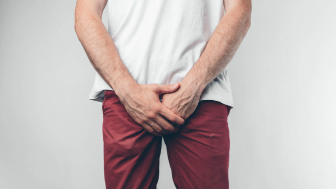 Caucasian man in white T-shirt and burgundy pants. Holds his hands on groin.