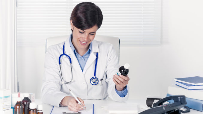 Smiling young doctor writing out a prescription for a bottle of medication as she sits in her office at the hospital