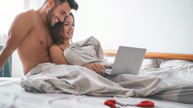 Happy couple watching on computer while lying on bed under blanket