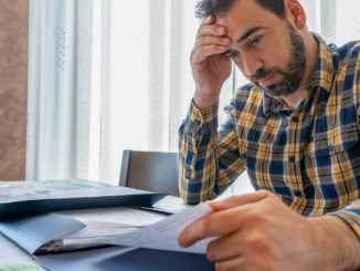 Bearded man dressed in plaid shirt looking at bills worried in his living room.