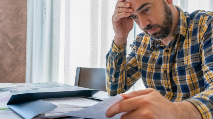 Bearded man dressed in plaid shirt looking at bills worried in his living room.