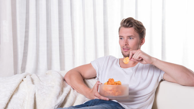 Young man sitting on couch and eating chips.