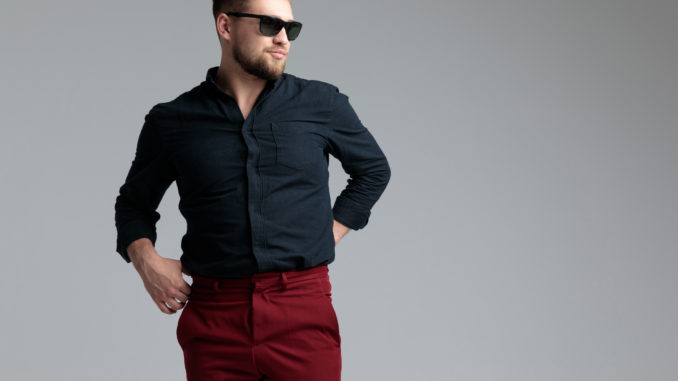 Detemined fashion man adjusitng his pants and looking away while wearing sunglasses, a black shirt and red jeans, standing on gray studio background