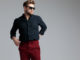 Detemined fashion man adjusitng his pants and looking away while wearing sunglasses, a black shirt and red jeans, standing on gray studio background