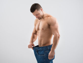 Mid adult muscular man looking at weight loss while holding old jeans against white background