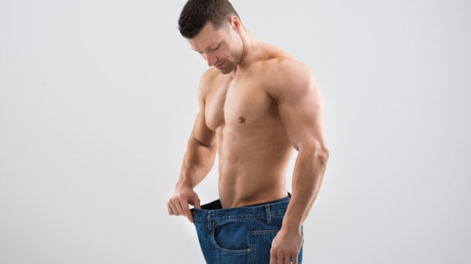 Mid adult muscular man looking at weight loss while holding old jeans against white background