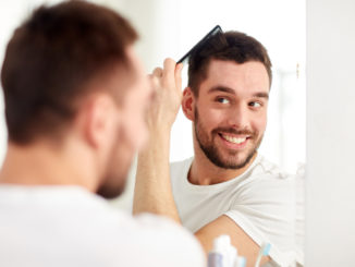 Beauty, grooming and people concept - smiling young man looking to mirror and brushing hair with comb at home bathroom