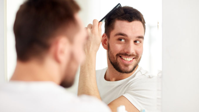 Beauty, grooming and people concept - smiling young man looking to mirror and brushing hair with comb at home bathroom