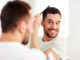 Beauty, grooming and people concept - smiling young man looking to mirror and brushing hair with comb at home bathroom