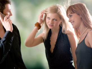 Young man speaking by mobile phone and two women looking at him and smiling, outdoors, focus on woman with red hair and man