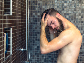 Young man with a beard standing washing his hair in the shower with shampoo rinsing it off under the jet of water, upper body with tattoo