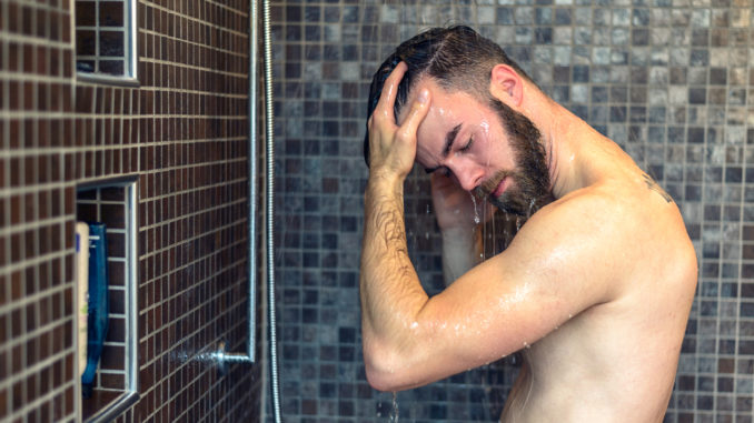 Young man with a beard standing washing his hair in the shower with shampoo rinsing it off under the jet of water, upper body with tattoo