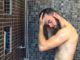 Young man with a beard standing washing his hair in the shower with shampoo rinsing it off under the jet of water, upper body with tattoo