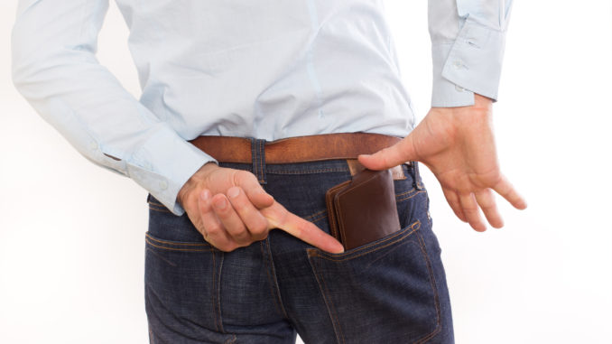 Attractive young man in jeans and shirt putting leather brown wallet in back pocket, against white background