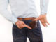 Attractive young man in jeans and shirt putting leather brown wallet in back pocket, against white background