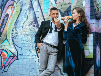 couple in luxury clothing eating hot dogs near wall with graffiti on street