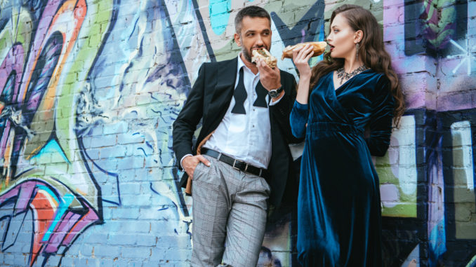 couple in luxury clothing eating hot dogs near wall with graffiti on street