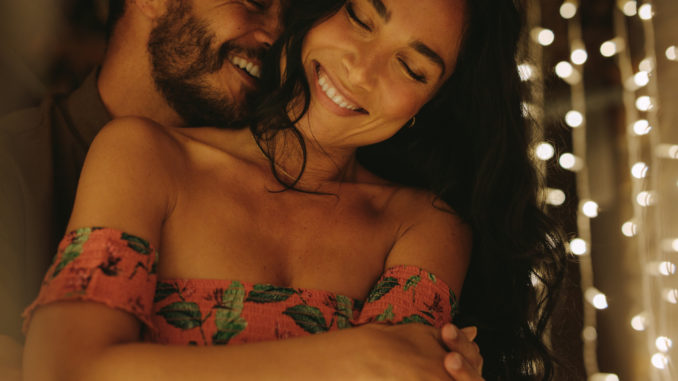 Romantic couple embracing and enjoying an intimate moment together, against the backdrop of lights. Handsome men embracing his beautiful girlfriend from behind