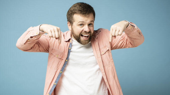 Charming, happy bearded man looks and points his index finger down, smiling broadly and looking at the camera. Isolated on a blue background.