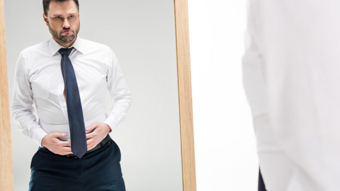 Dissatisfied chubby man in formal wear looking at mirror on white with copy space