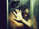 Young couple of naked women and men taking shower with water drops embracing in passion with wet bodies with hand on glass