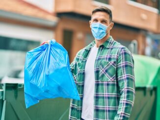 Young caucasian man wearing medical mask throwing waste bag to the container at the city