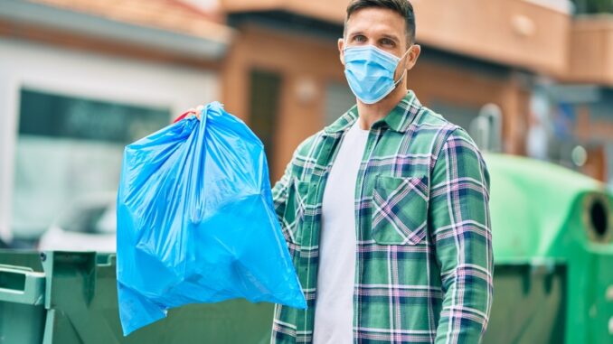 Young caucasian man wearing medical mask throwing waste bag to the container at the city