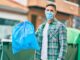 Young caucasian man wearing medical mask throwing waste bag to the container at the city