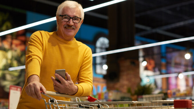 Portrait of modern senior man grocery shopping in supermarket pulling shopping cart and smiling