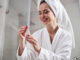 Excited young woman wearing bathrobe and towel on her head holding and looking at small bottle with cosmetic product