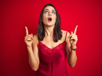 Young beautiful woman wearing lingerie over red isolated background amazed and surprised looking up and pointing with fingers and raised arms