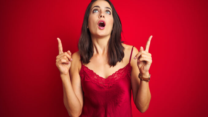 Young beautiful woman wearing lingerie over red isolated background amazed and surprised looking up and pointing with fingers and raised arms