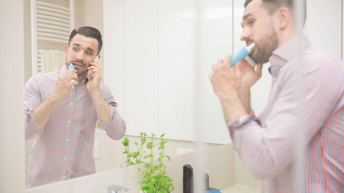 Man standing in bathroom, brushing teeth and having phone call.